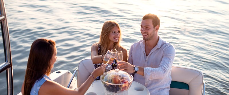 section 3-img Three people toasting with drinks on a boat during sunset, sitting around a table with fruit.