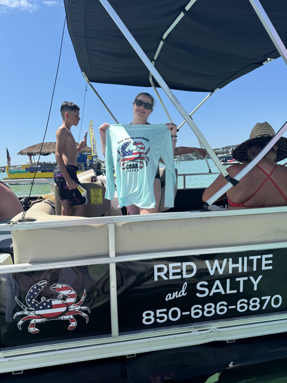 People on a boat, one holding a shirt with a crab design, under a canopy on a sunny day.